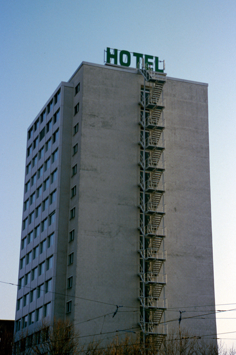 a tall, simple building with green letters spelling ‚hotel' at the top. a narrow fire escape runs down the pale concrete wall from the hotel sign to the ground.