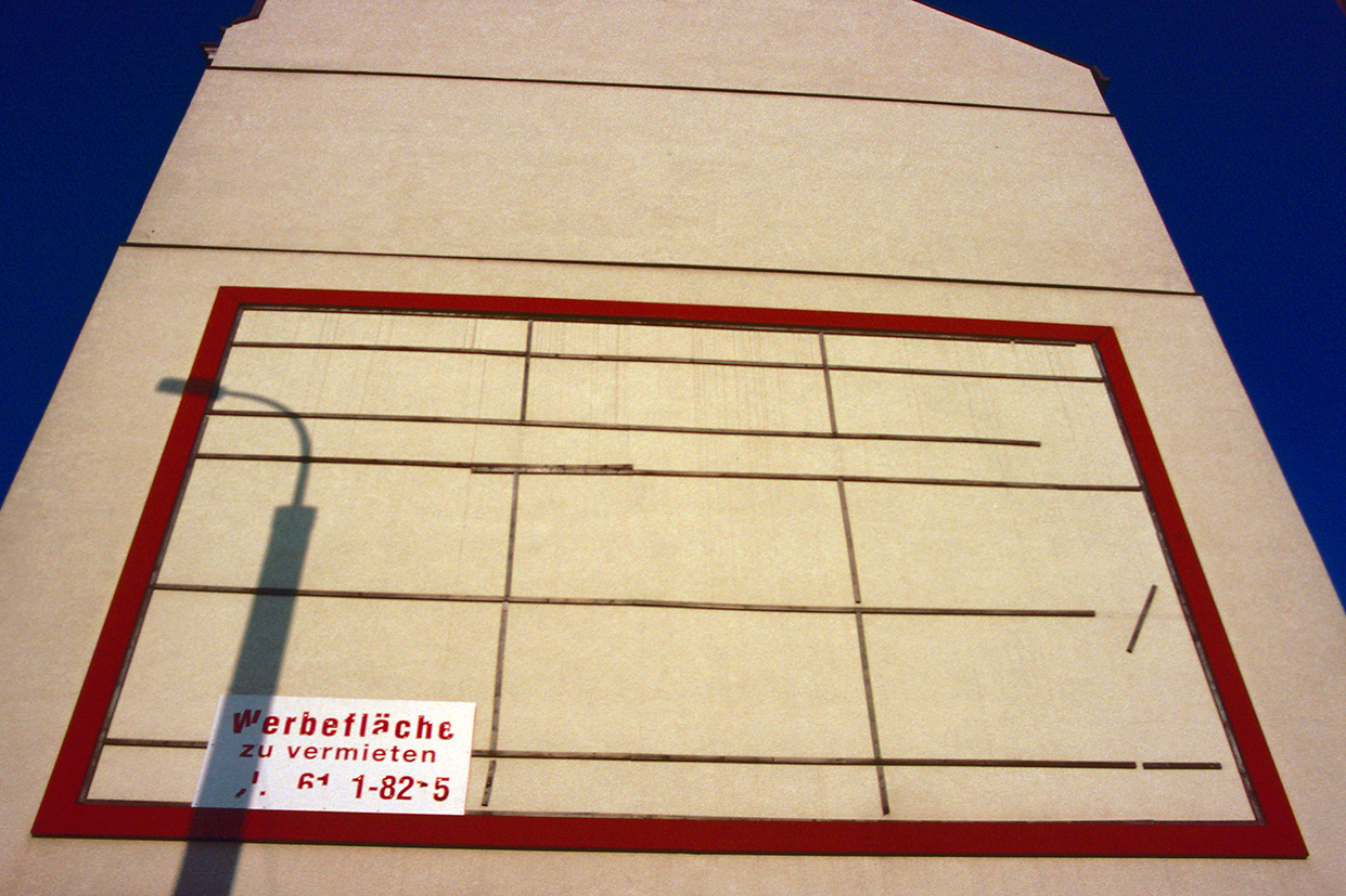 upward view of a bare, windowless wall against a blue sky, with a red empty advertising frame and the shadow of a streetlight.